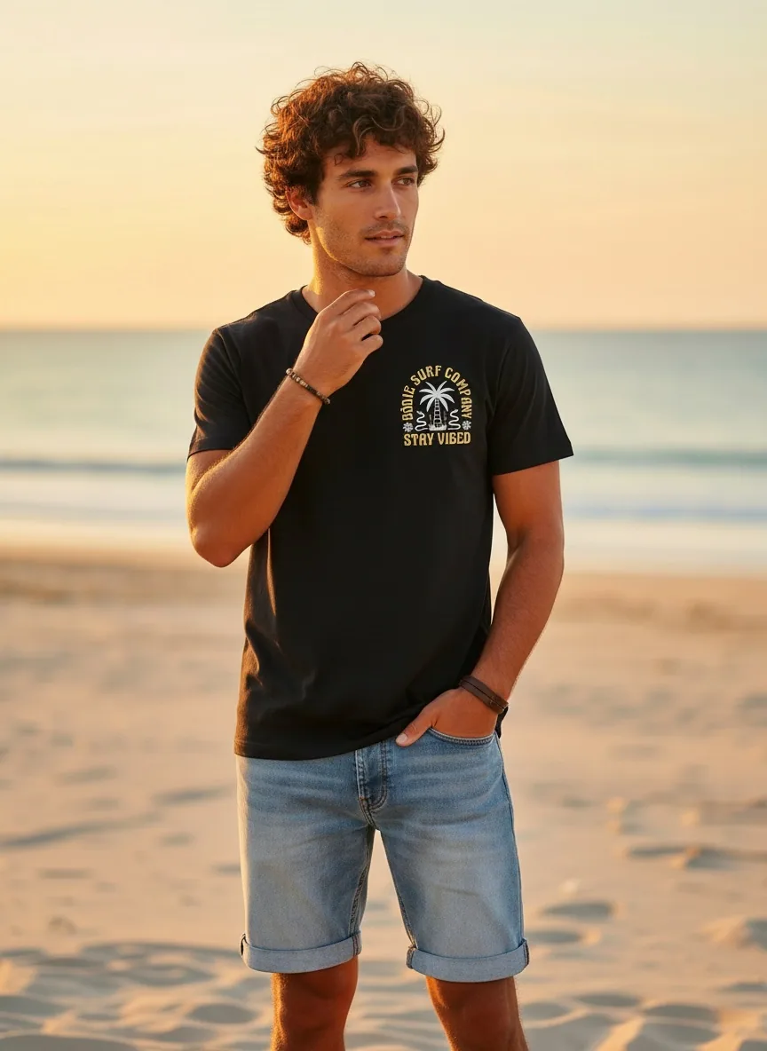 Bōdie Surf Co black STAY VIBED tee front with chest logo on AI male model on Gulf Coast beach at golden hour